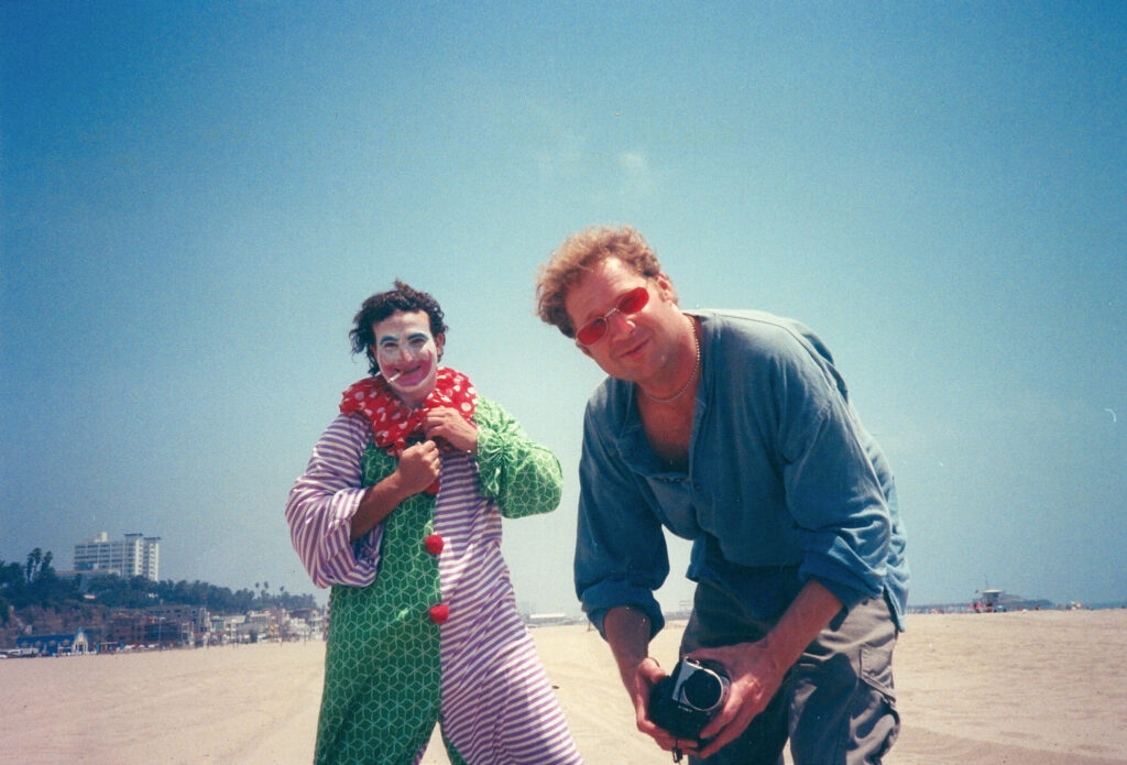 A man dressed as a clown with a cigarette dangling from his mouth looks into the camera along with a man in rose colored glasses holding a camera in this behind the scenes photo taken at Santa Monica beach.